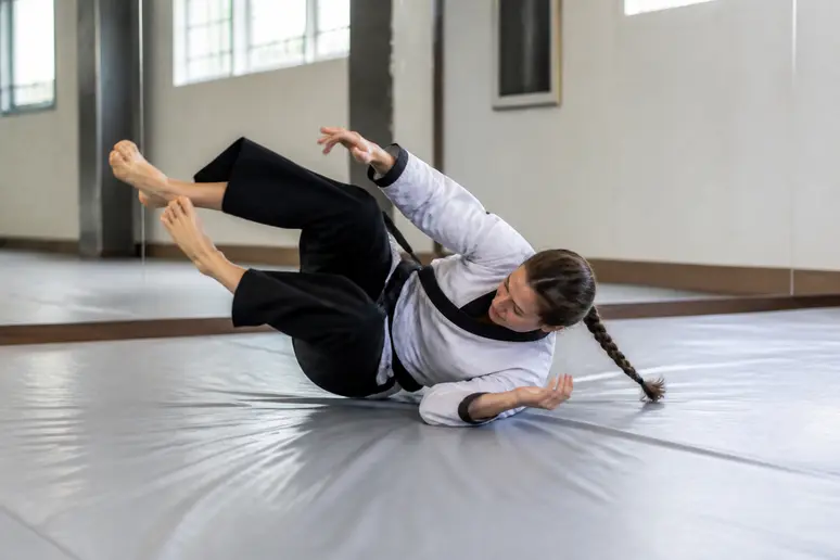 Personne en uniforme pratiquant une roulade de chute sécurisée sur un tatami.